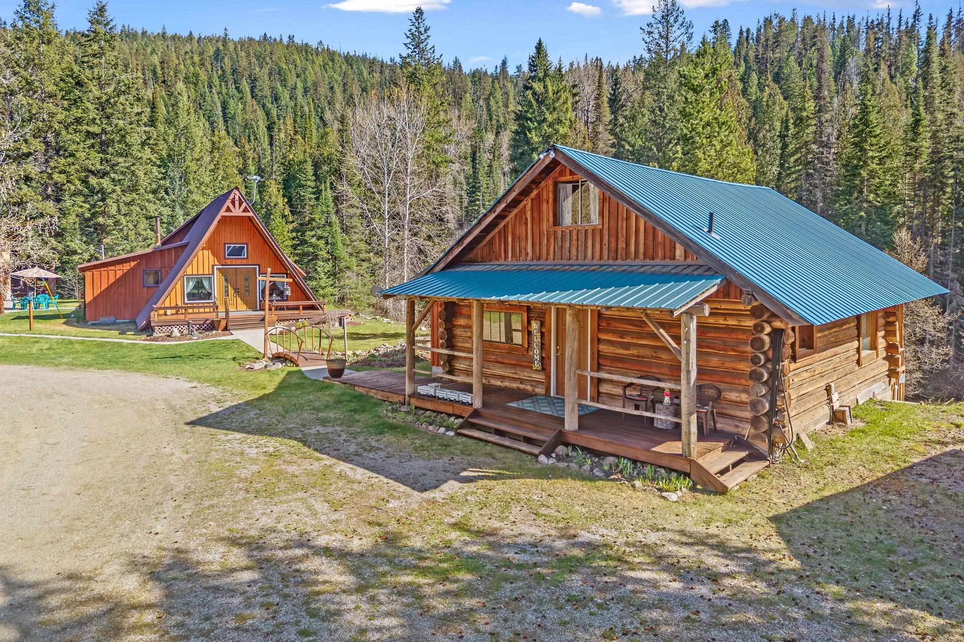 Two log cabins with blue roofs in a forest clearing under a bright sky.