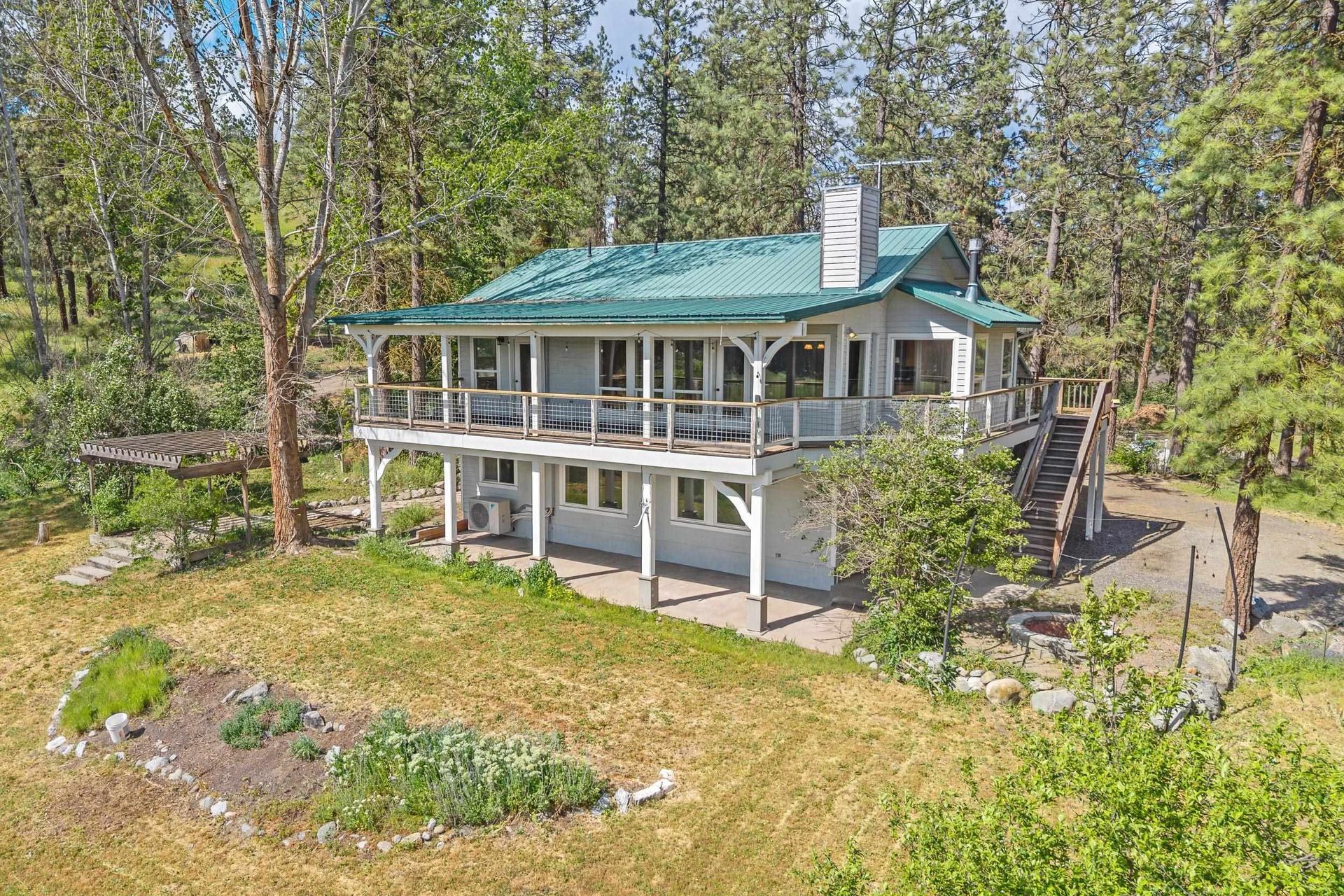 Two-story cabin with teal roof and wraparound porch in a wooded clearing