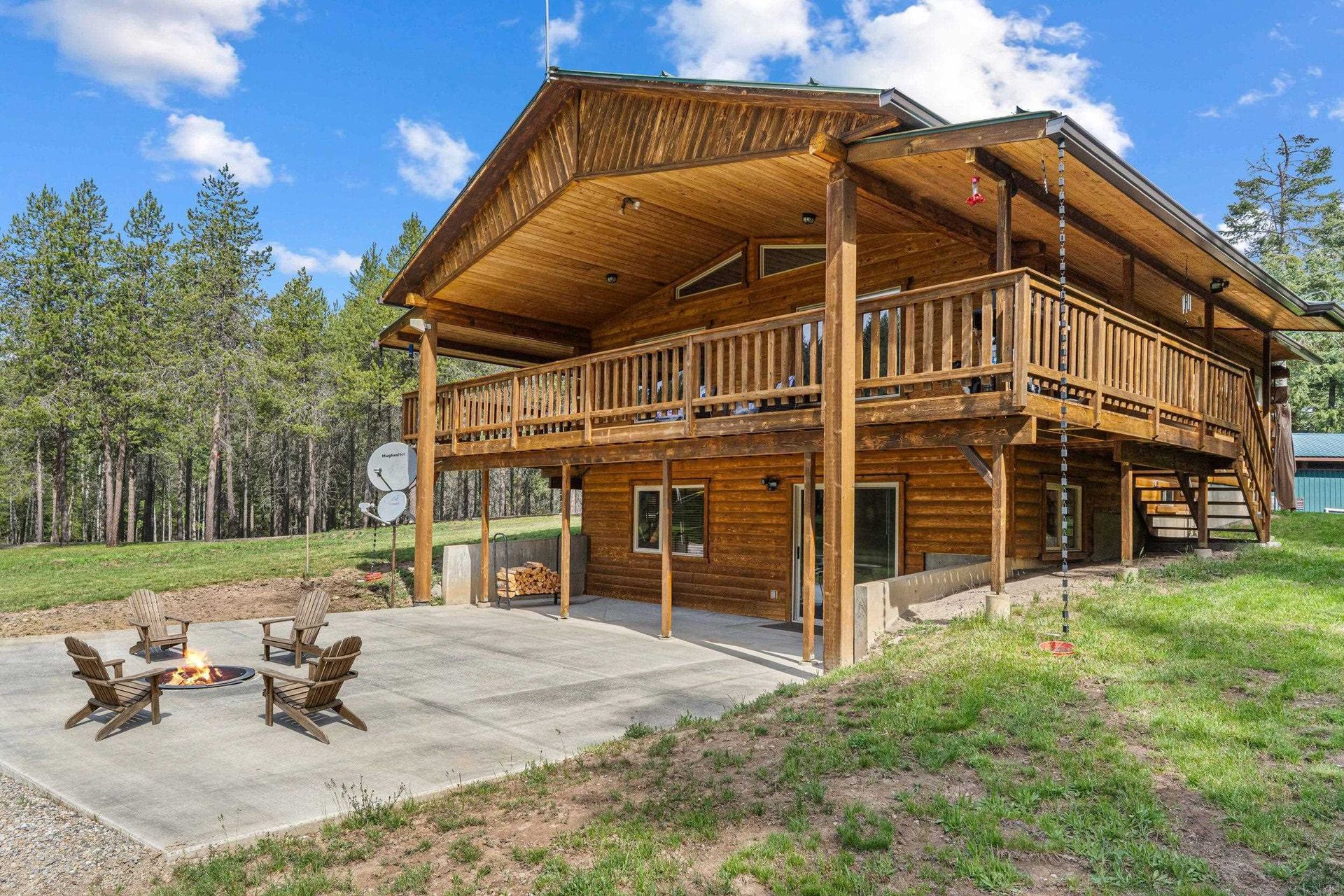 Rustic wooden cabin with wraparound porch and two chairs on a concrete patio, surrounded by trees and grass.