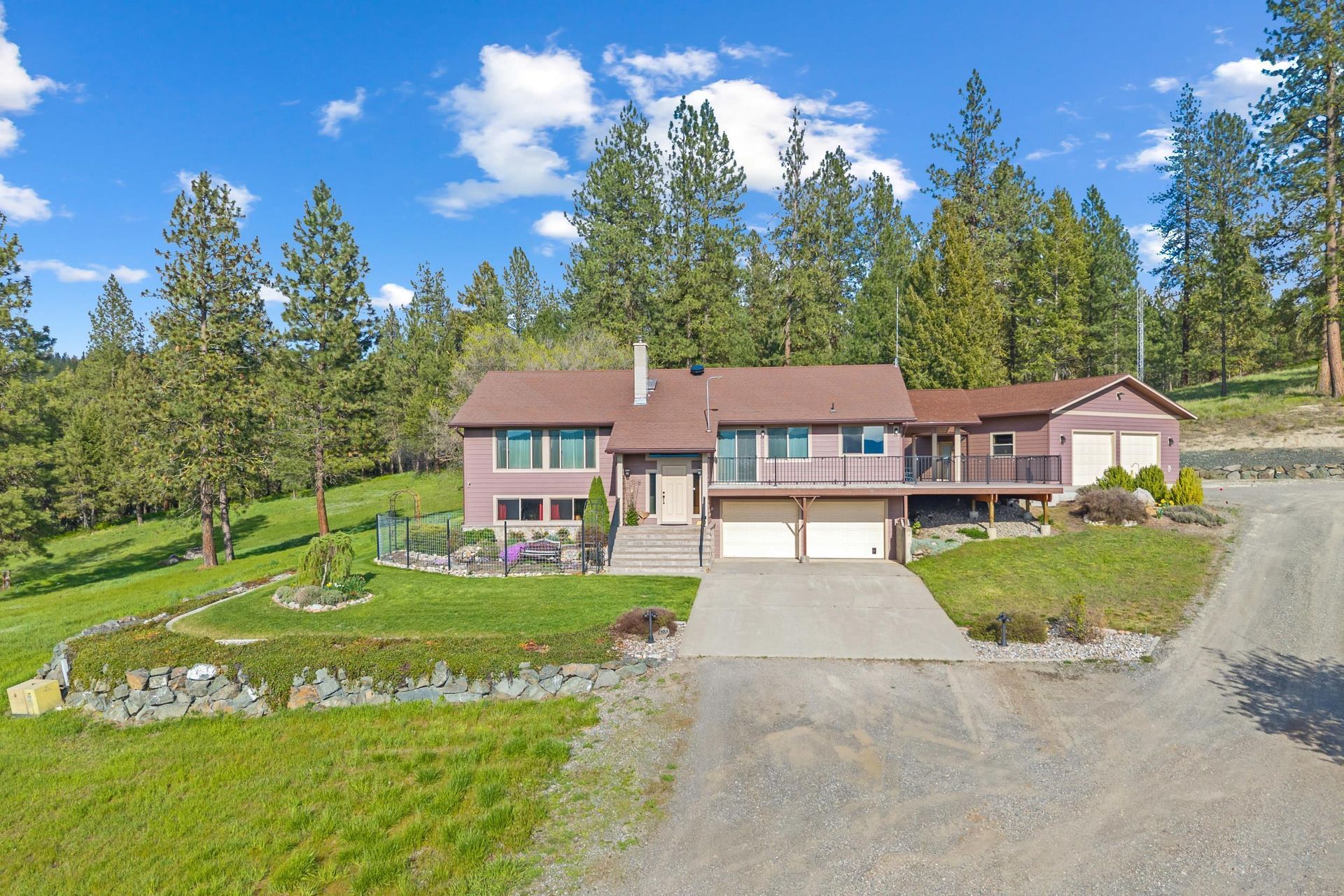 Two-story brown house with a driveway, garage, and wooded hillside under a blue sky