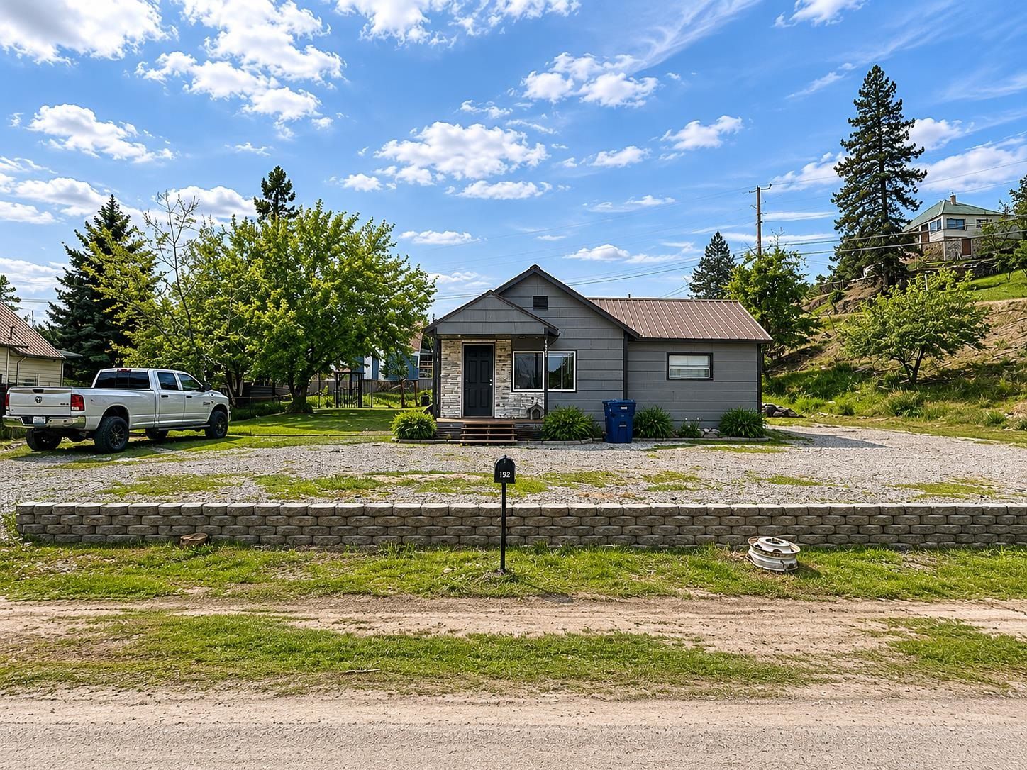 Small gray house with a gravel yard, parked white truck, trees, and a bright blue sky with clouds