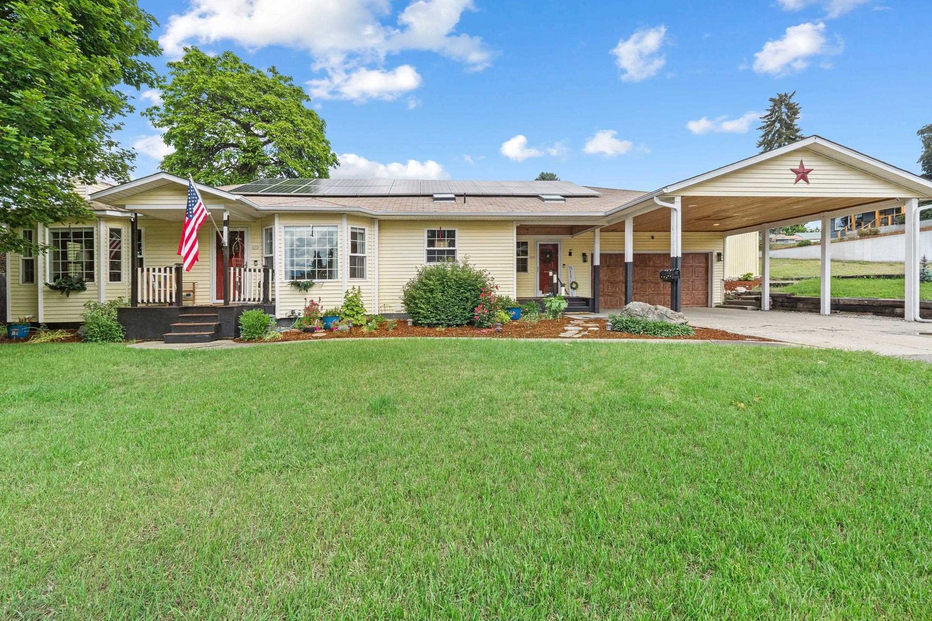 Single-story white house with an American flag, green lawn, and attached carport under a blue sky