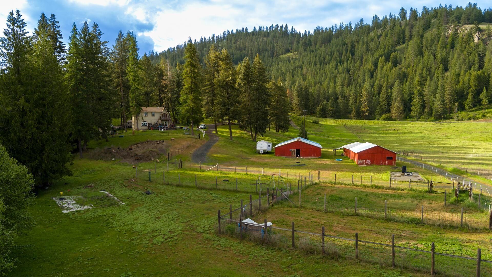 Green rural valley with red barns, a small house, fenced pasture, and forested hills under blue sky