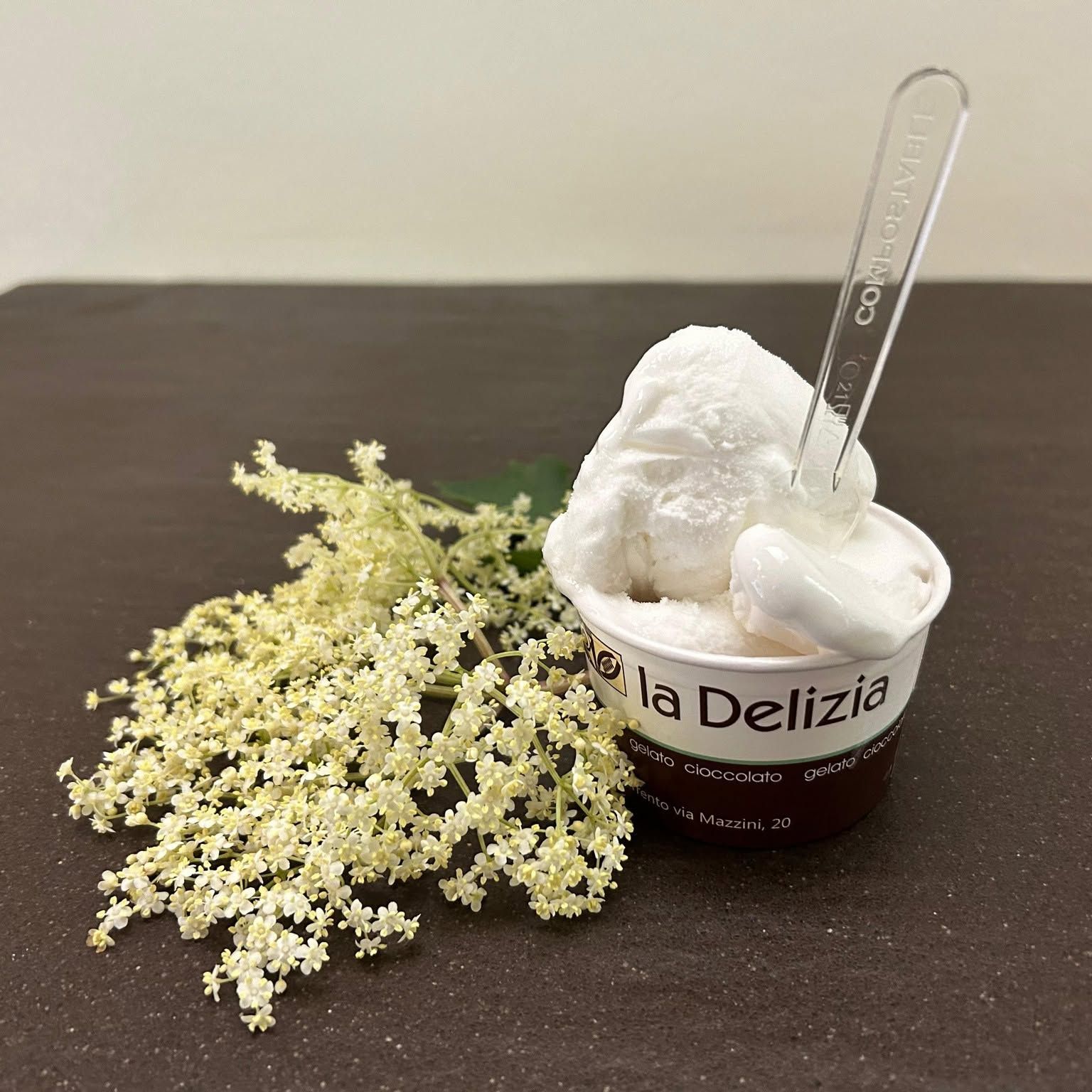 Coppetta di gelato con palline bianche, cucchiaio trasparente e fiori giallo pallido su un tavolo scuro.
