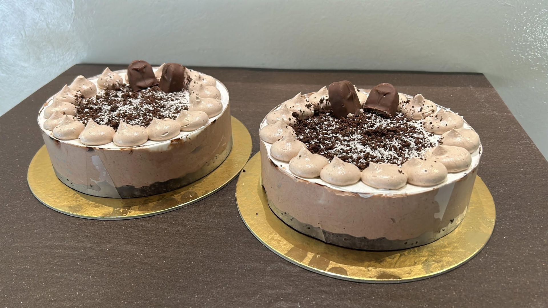 Due torte al cioccolato su vassoi dorati, decorate con ciuffi di panna e scaglie di cioccolato, adagiate su un tavolo scuro.