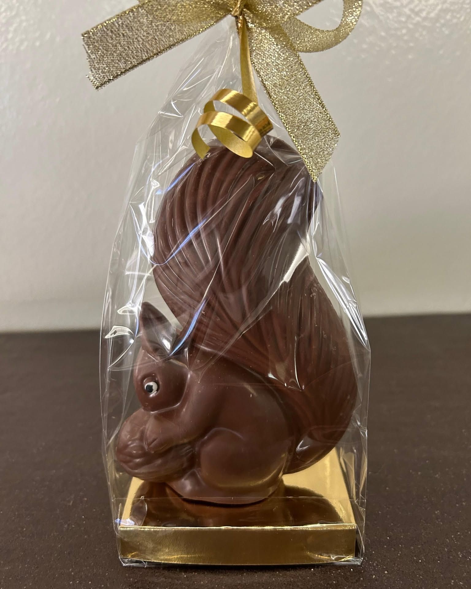 Coniglietto di cioccolato incartato in carta trasparente con nastro dorato, seduto su una base dorata su sfondo neutro.