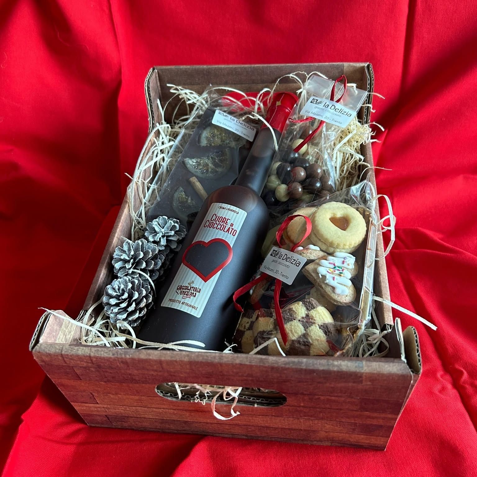 Cesto regalo con vino in scatola di legno, bottiglia, decorazione a forma di pigna, frutta secca e biscotti su tessuto rosso.