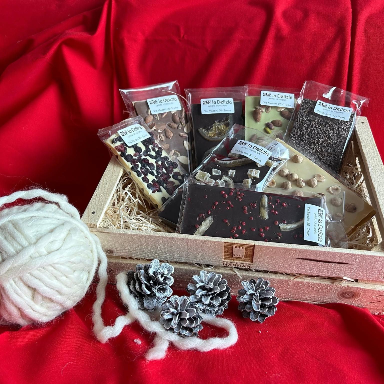 Tavoletta di cioccolato assortita in una scatola rosa su tessuto rosso, con filo bianco e pigne glassate nelle vicinanze.