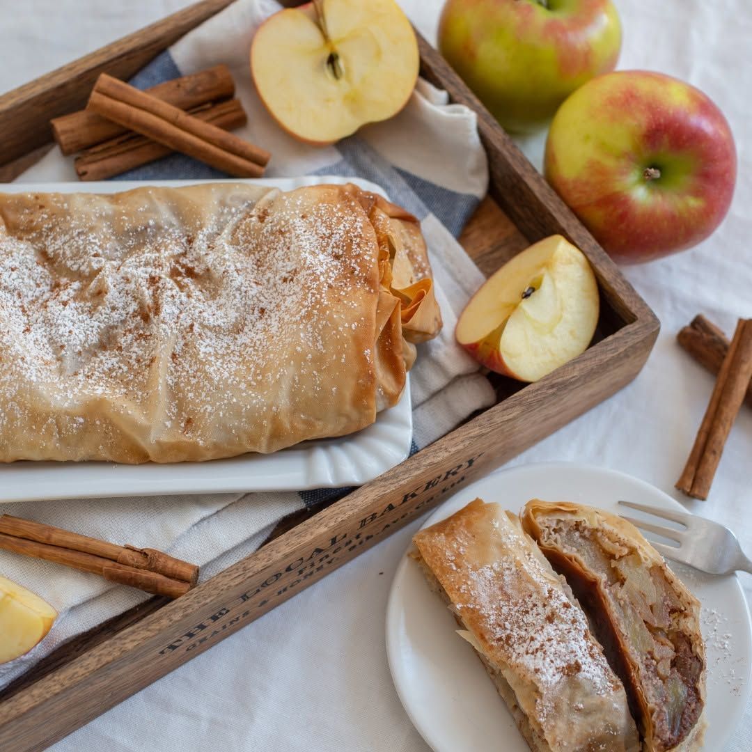 Strudel di mele spolverato di zucchero a velo su un vassoio, con bastoncini di cannella e mele a fette accanto.