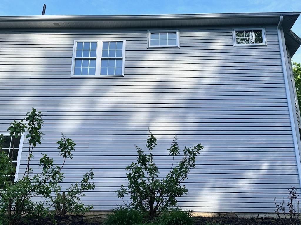 Gray vinyl-sided house with three windows under a blue sky; shrubs in foreground.