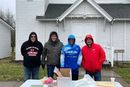 Four people stand behind a table outside a white church. They wear winter jackets, and some have hoods up.
