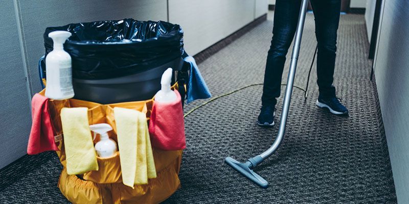 Person vacuuming a carpeted hallway, cleaning supplies in a cart nearby.