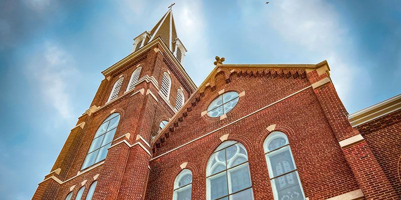 Low-angle shot of a brick church with a tall steeple against a cloudy sky.