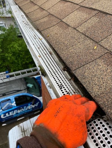 Person in orange glove cleaning a gutter covered with a screen on a roof near shingles.