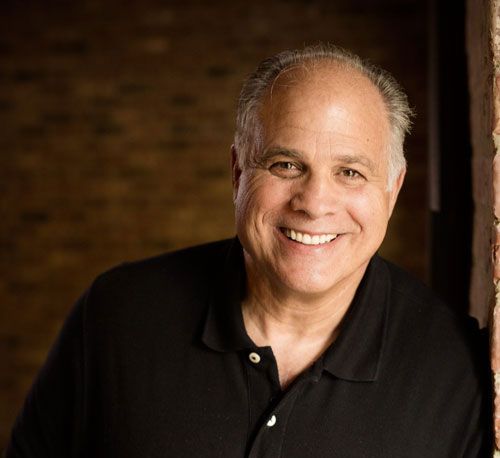 A man in a black shirt is smiling and leaning against a brick wall.