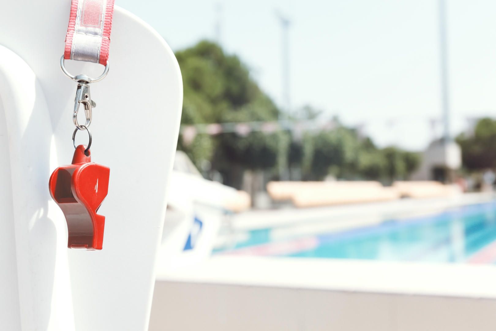 A red whistle is hanging from a white chair in front of a swimming pool.