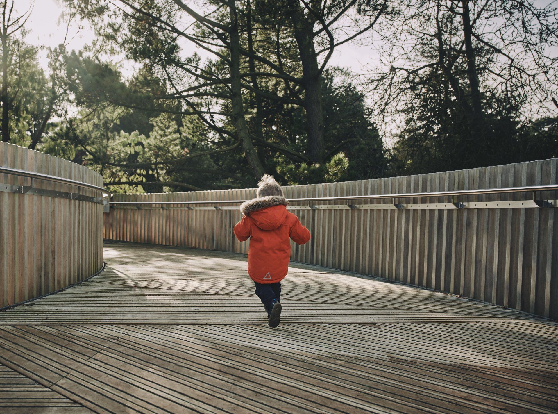 A little boy in an orange jacket is running across a wooden bridge.