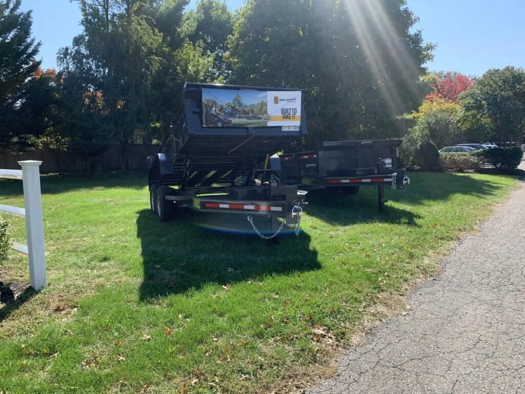Trailer with sign on grass, near a paved path, in front of trees under sunny sky. | Dave Muller Automotive & Trailer