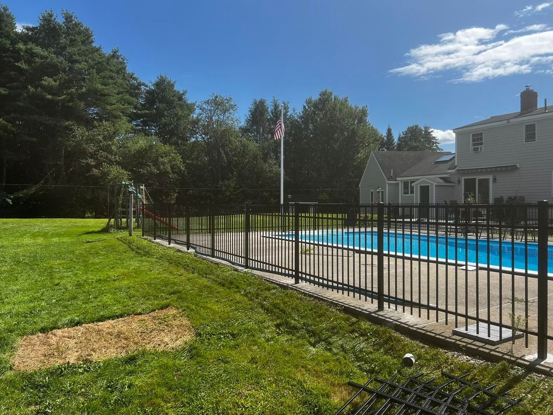 A backyard with a pool surrounded by a black fence. Trees and a house are in the background.