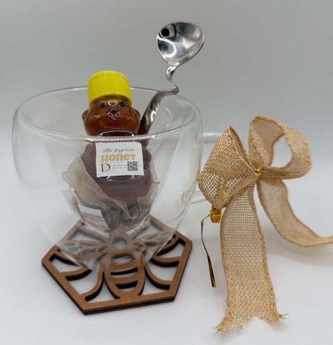 Heart-shaped glass tea cup holding honey bear and tea bags, displayed on wooden bee coaster with bow.