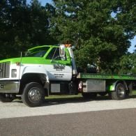 Green and white tow truck parked on pavement near trees.