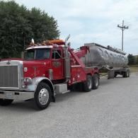 Red tow truck hauling a large silver tank trailer on a paved lot.