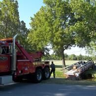 Red tow truck pulling a trailer off the side of a road; a man stands nearby, trees in the background.