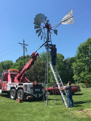 Two men repair a windmill with a tow truck assisting on a sunny day.