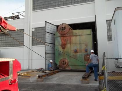 Crane pulling a large, green industrial machine out of a building. Two workers assist, wearing hard hats.