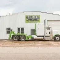 White and green semi-truck in front of Thomas 24/7 Towing & Recovery building.