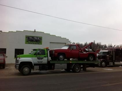 Tow truck towing a red pickup truck in front of a garage. The tow truck is green and white. A semi truck is visible.
