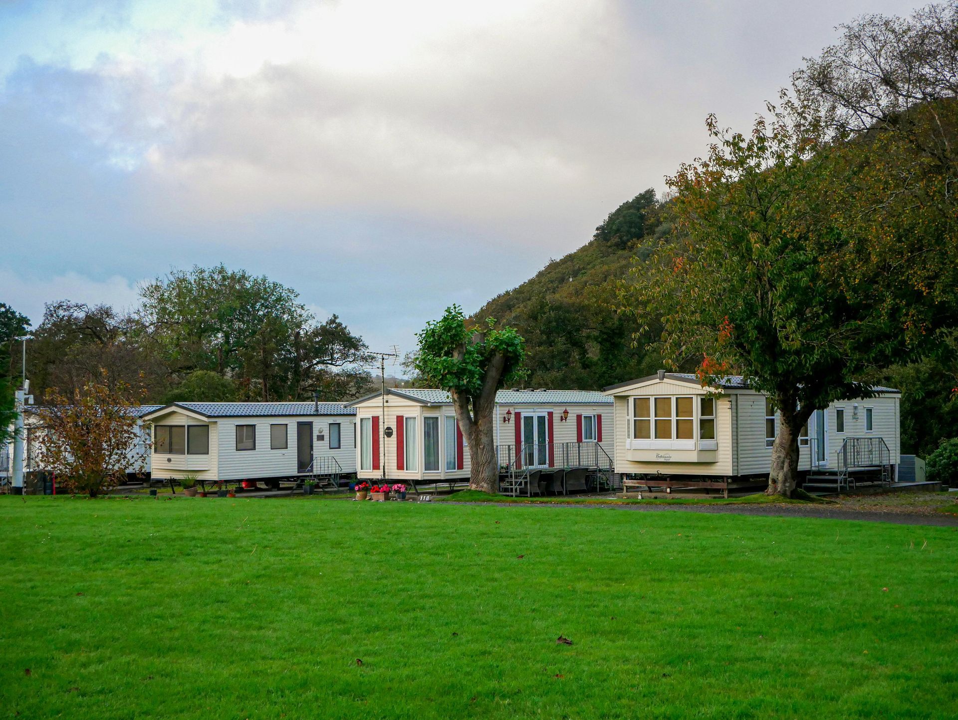 Row of white cabins with red and yellow trim beside a large green lawn and wooded hill under cloudy sky