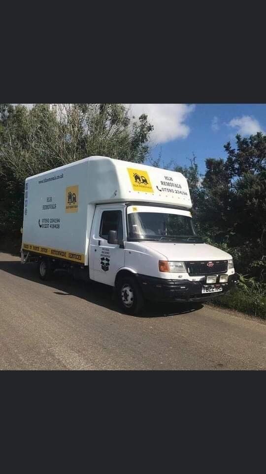 White moving truck with yellow logo parked on a roadside beside trees under a blue sky