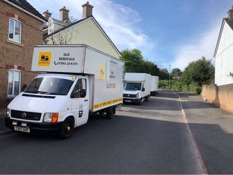Two white box trucks parked on a residential street, one near a house and one farther down the road.