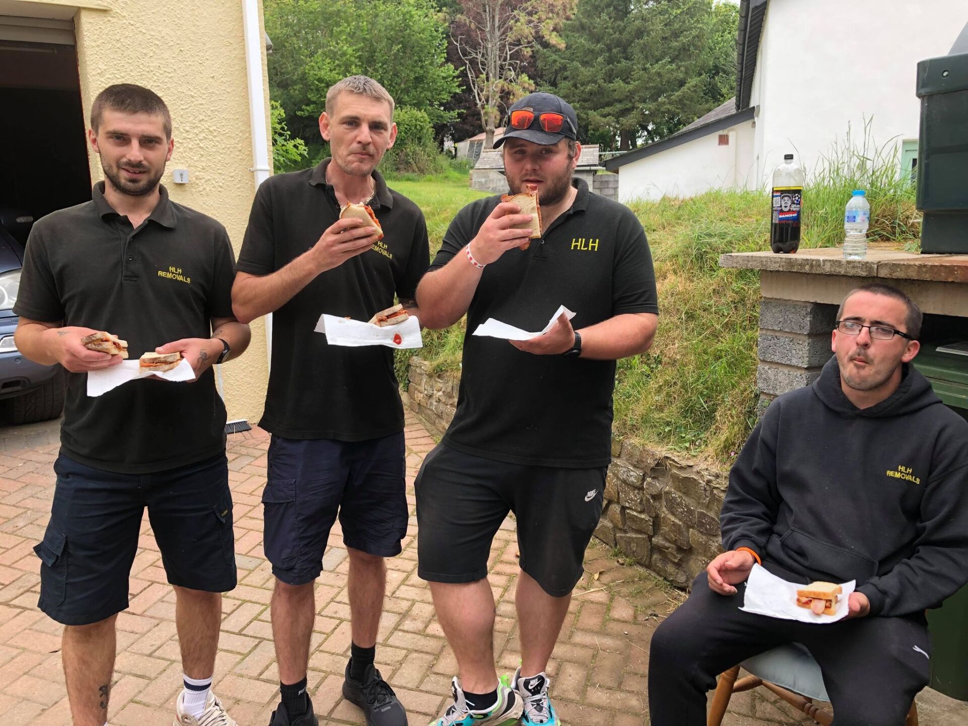 Four people in black sportswear eat from plates outdoors by a building and stone wall.