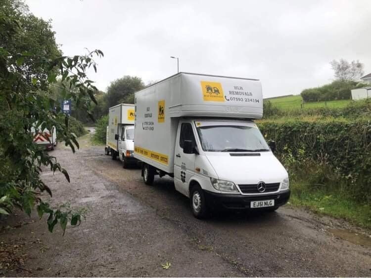 White moving truck on a narrow rural road beside green hedges on a cloudy day