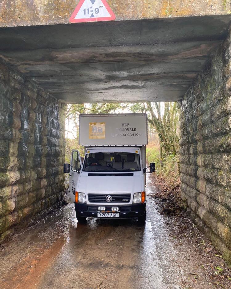 White delivery truck under a low stone bridge on a wet road, with a height warning sign above.