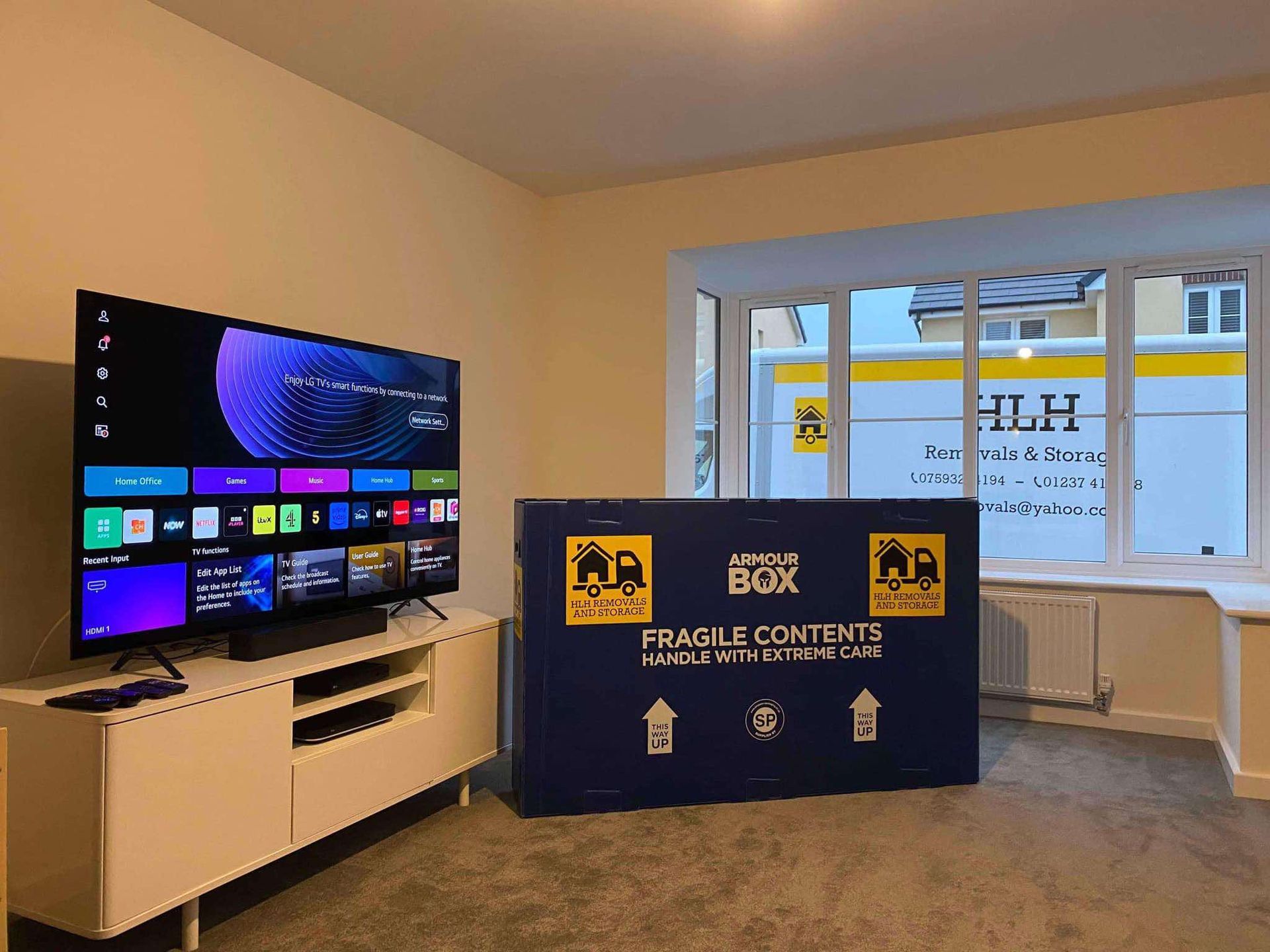 TV on stand beside blue donation box in a room with a window and street view
