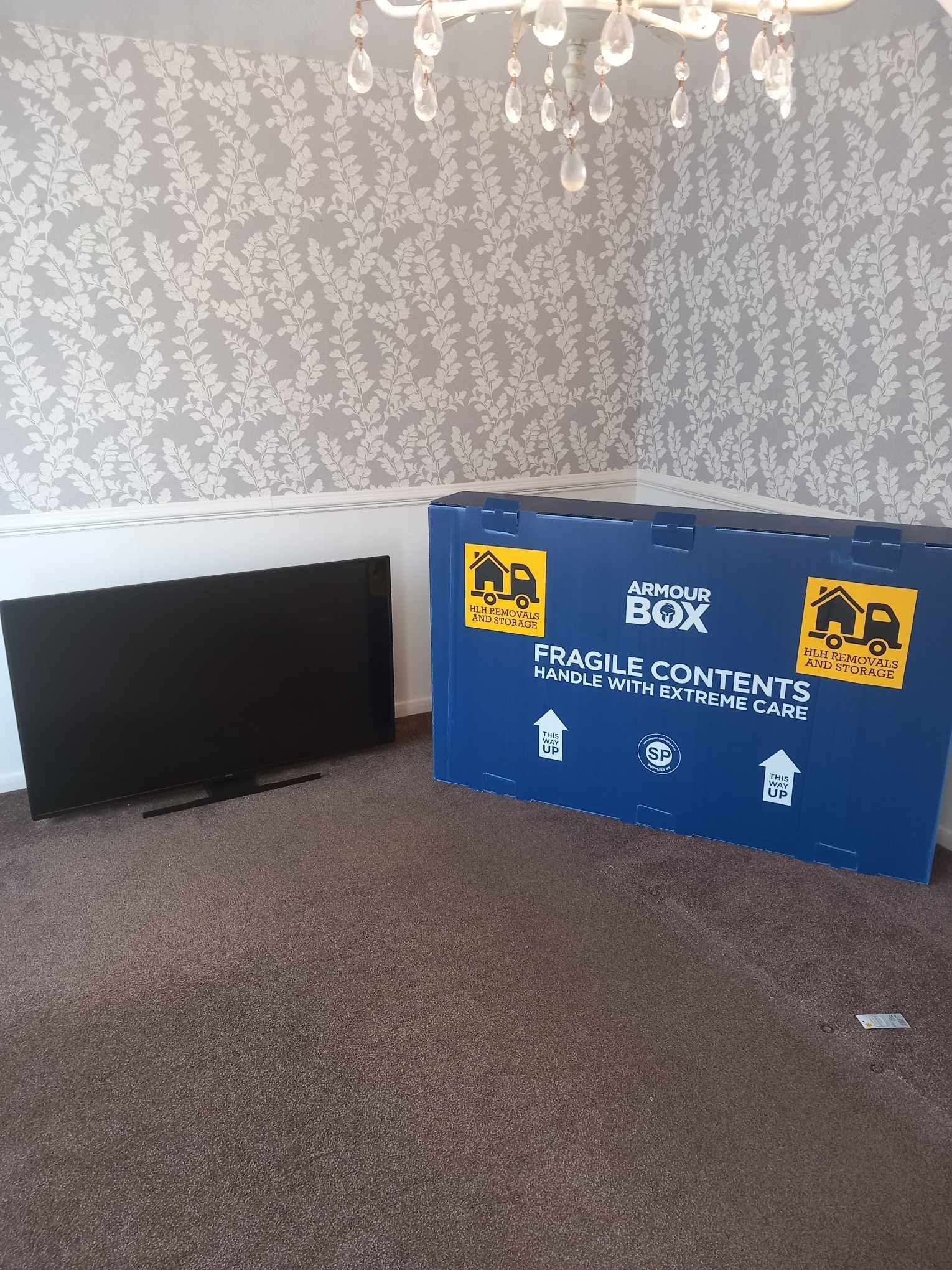 TV beside a large blue fragile shipping box in a carpeted room with patterned wallpaper