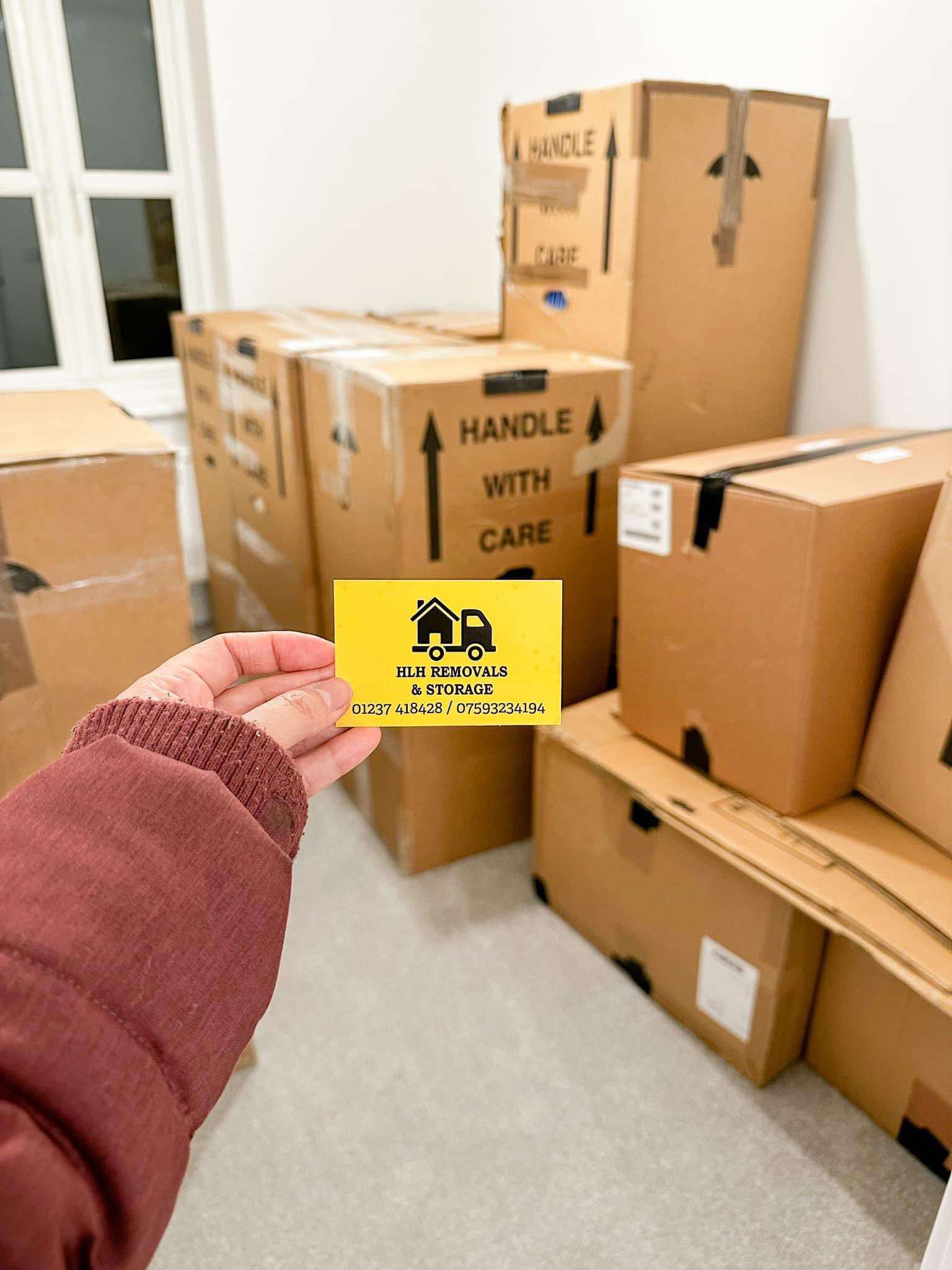 Hand holding a yellow card in front of stacked cardboard moving boxes in a room