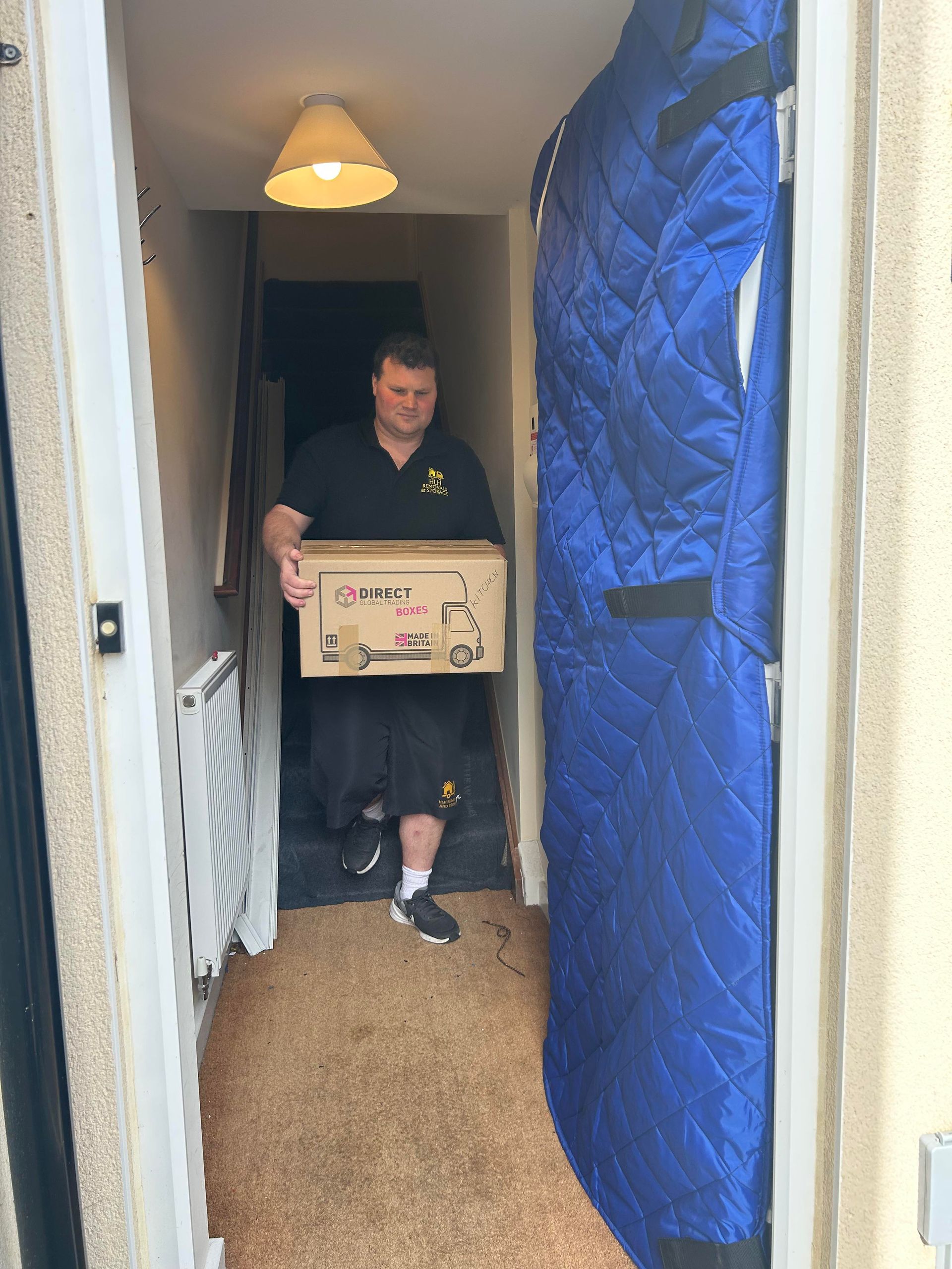 Man carrying a cardboard box through a narrow hallway with a blue moving blanket covering the right side.