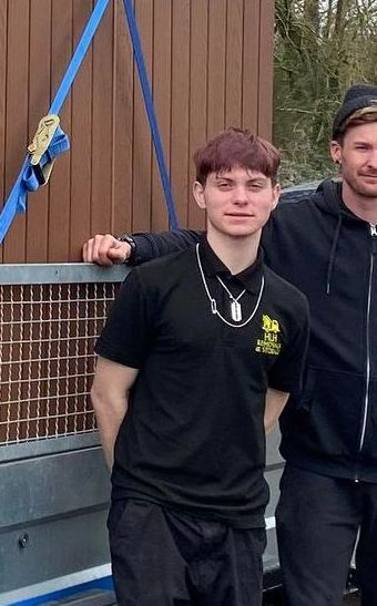 Two men standing outdoors beside a brown building, one in a black shirt leaning on a mesh barrier.
