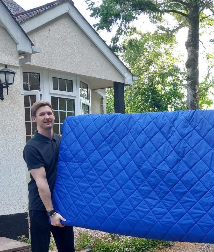 Person holding a large blue quilted mattress pad outdoors beside a house