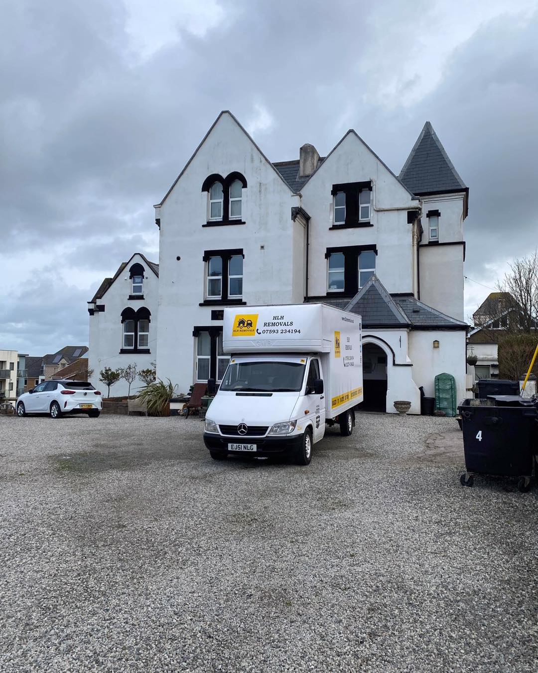 White multi-story house with two delivery vans parked on a gravel driveway under a cloudy sky