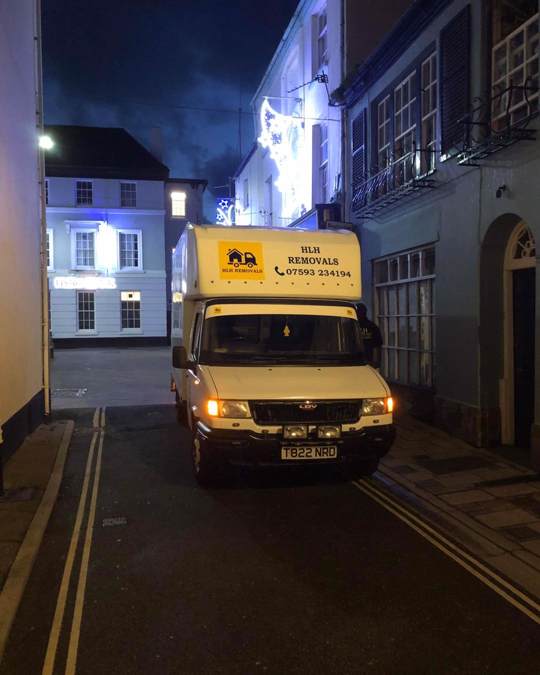 White van parked on a narrow night street beside lit buildings and a dark storefront