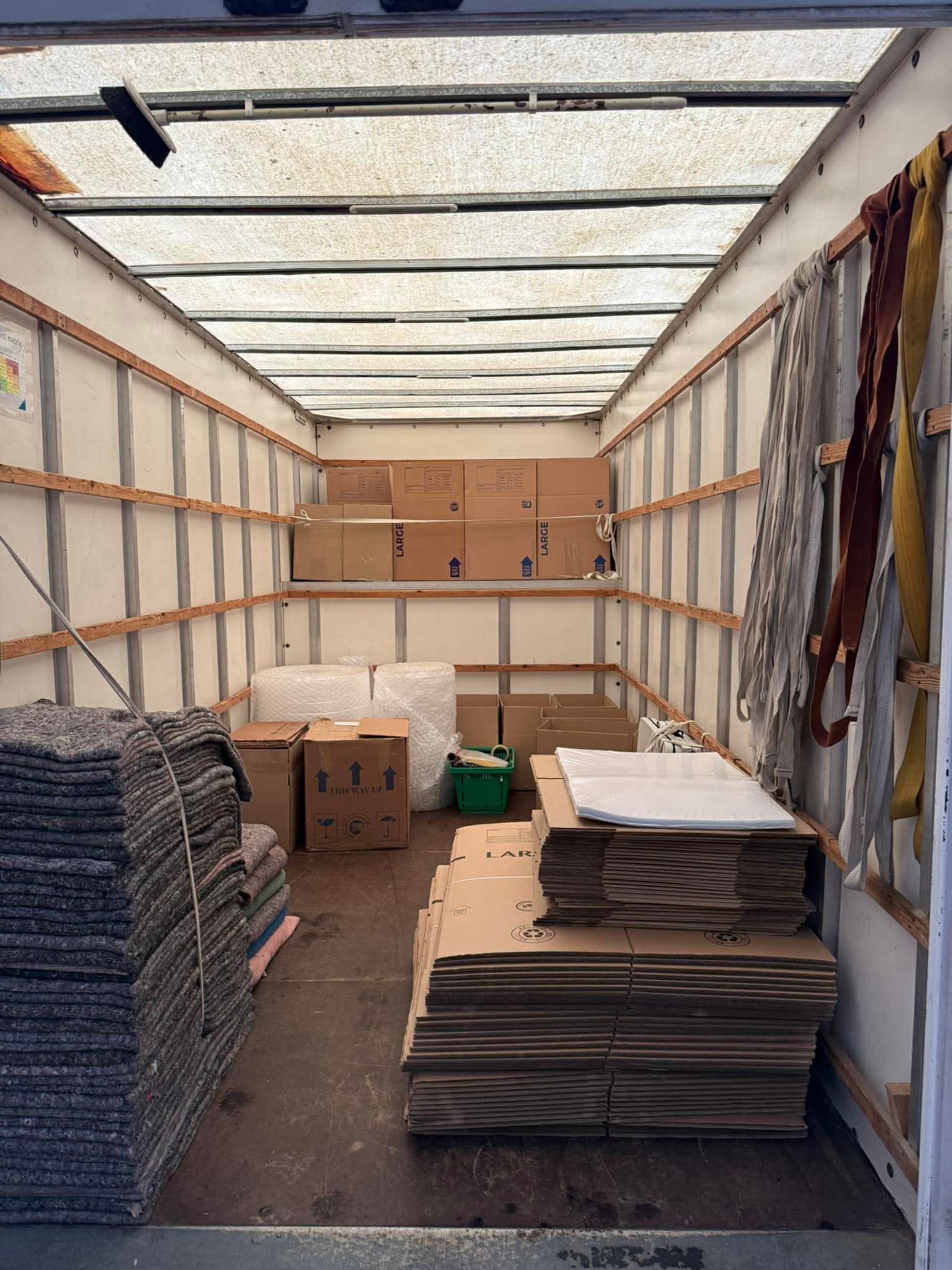 Inside a truck trailer, stacks of cardboard boxes and flat-packed materials are loaded for transport.