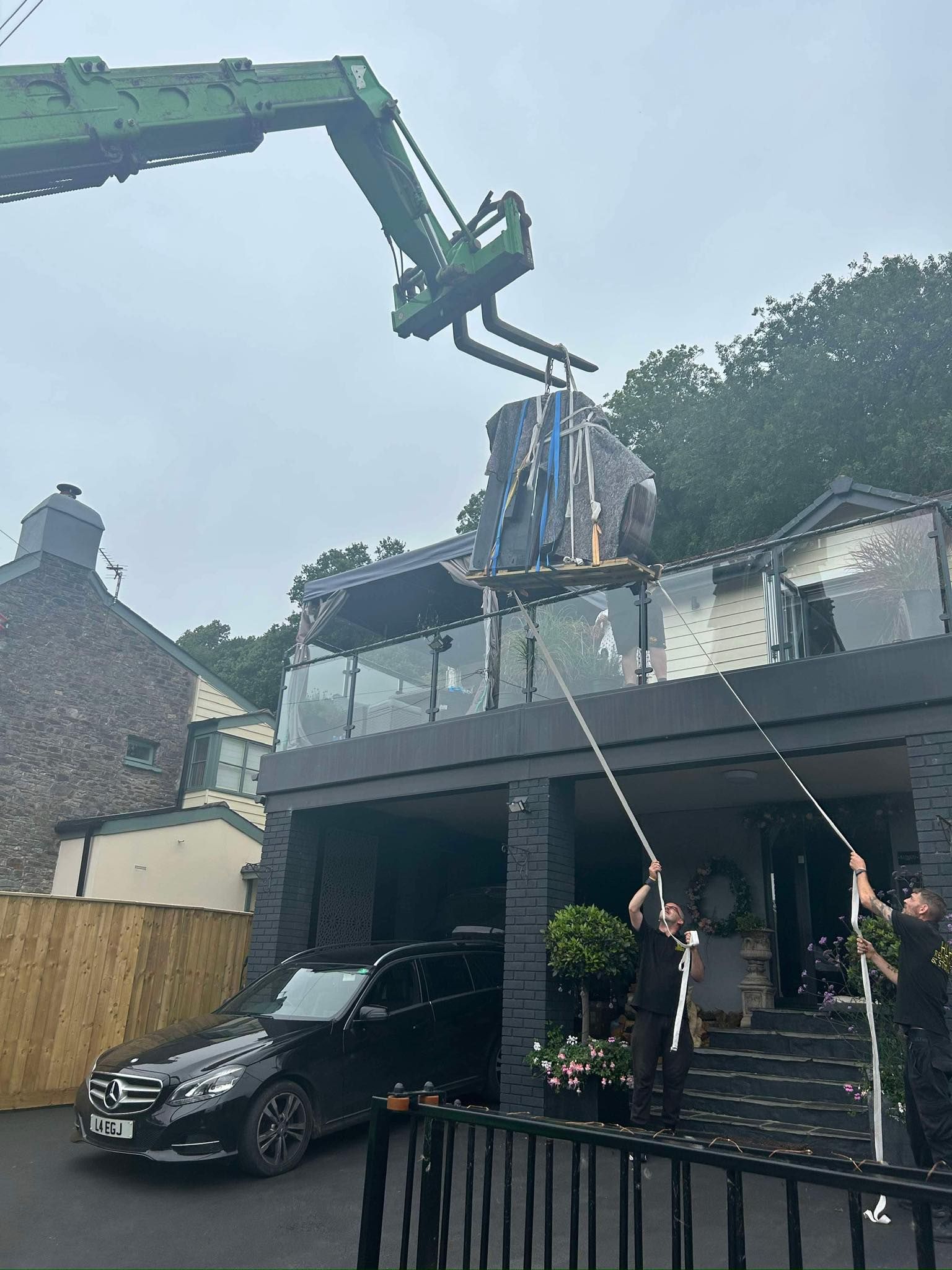Crane lifting materials onto a house roof while workers guide ropes in a residential driveway
