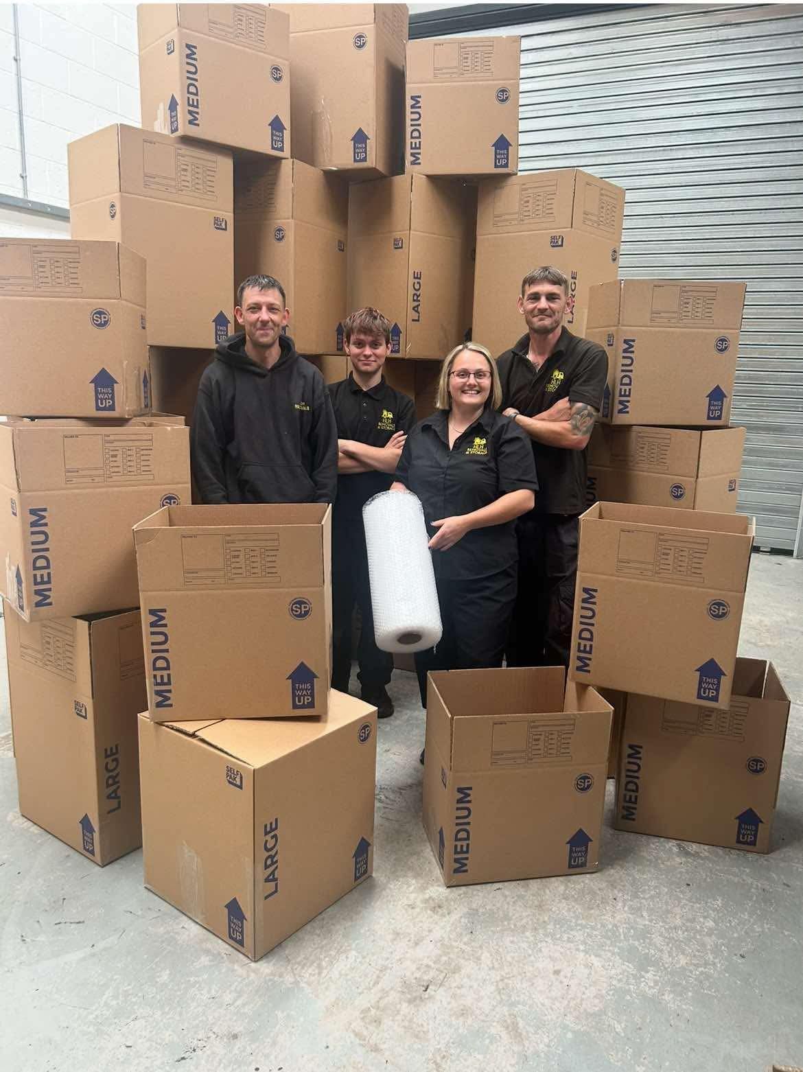 Four people stand smiling among stacked cardboard boxes in a warehouse, holding a roll of packing material.