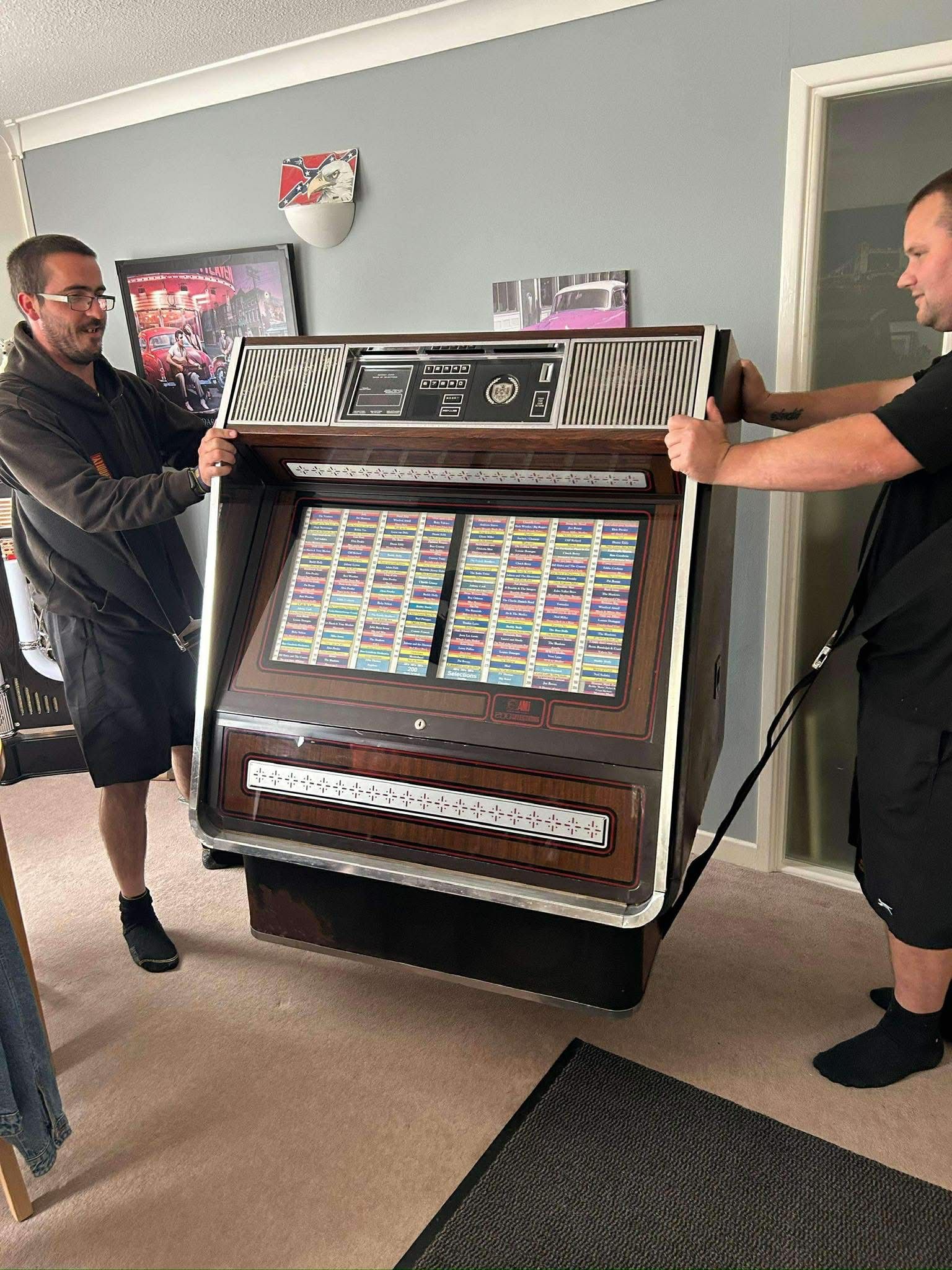 Two people carrying a large arcade-style jukebox in a room with gray walls and carpeted floor