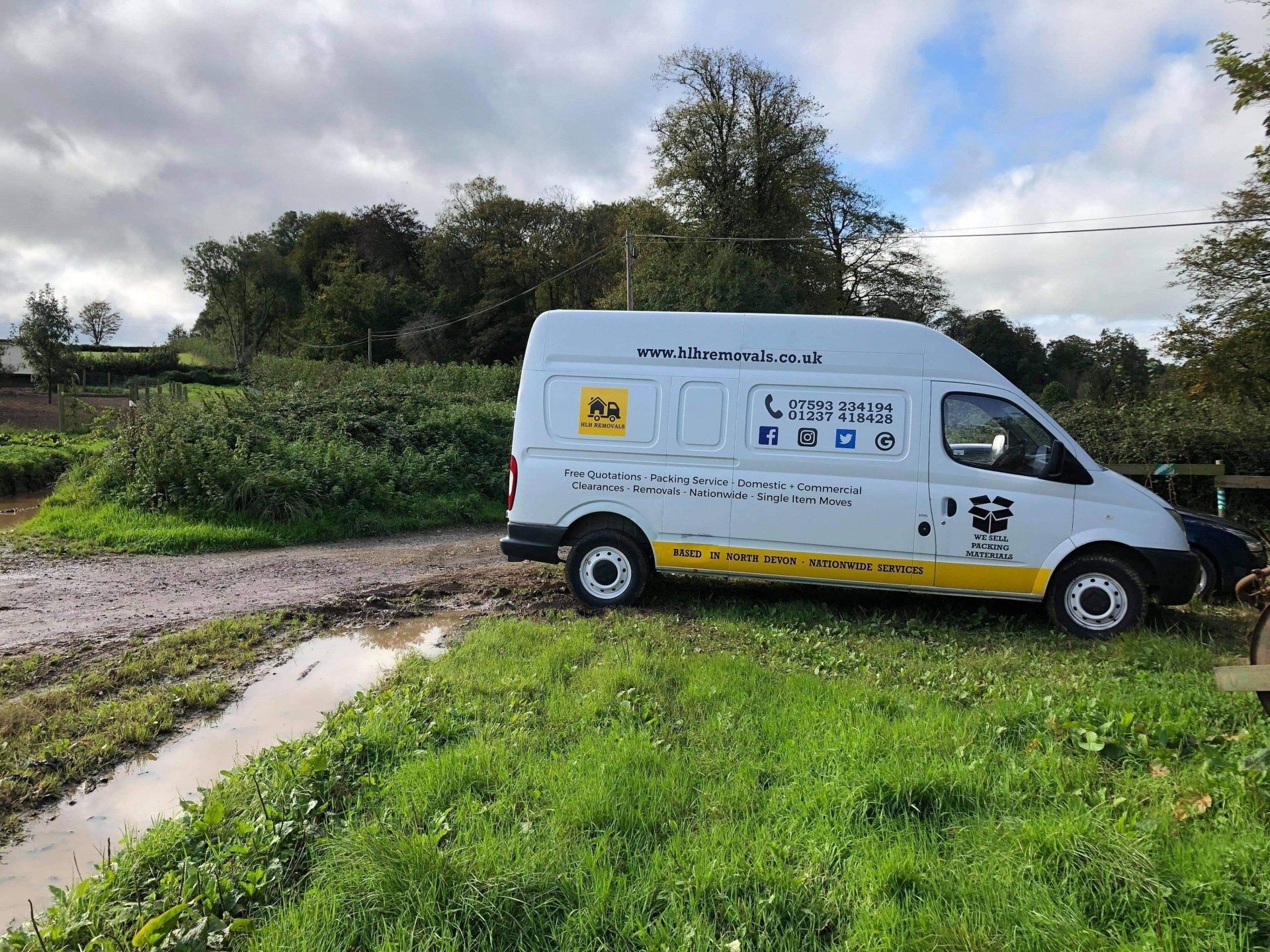 White delivery van parked on a dirt road beside a grassy field and trees under cloudy skies